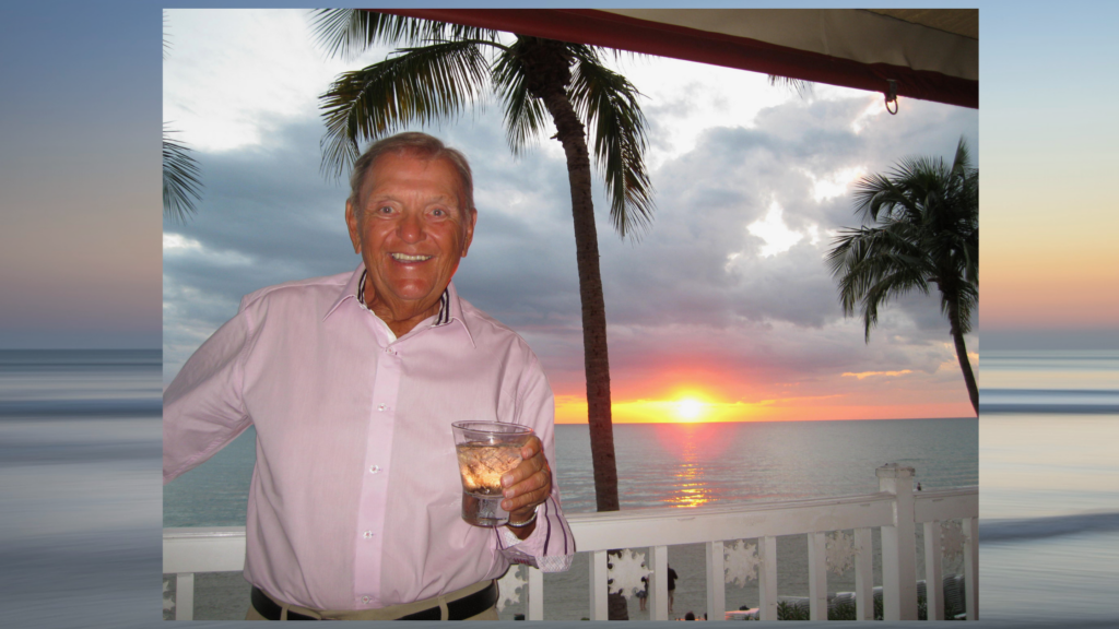 Smiling man in a pink shirt holding a drink on a balcony at sunset with palm trees and ocean in the background.