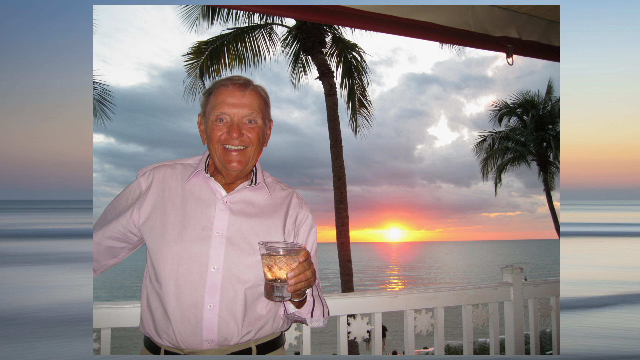 Smiling man in a pink shirt holding a drink on a balcony at sunset with palm trees and ocean in the background.